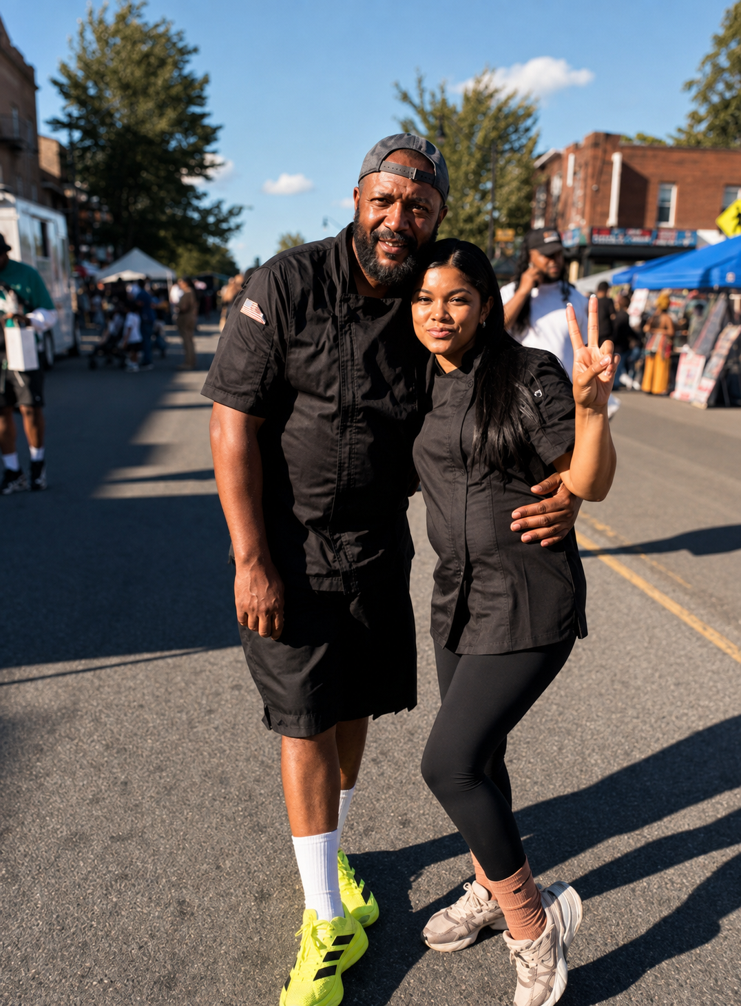 Fahkir and Nadia in black chef shirts standing together in the middle of a sunlit Newark street festival
