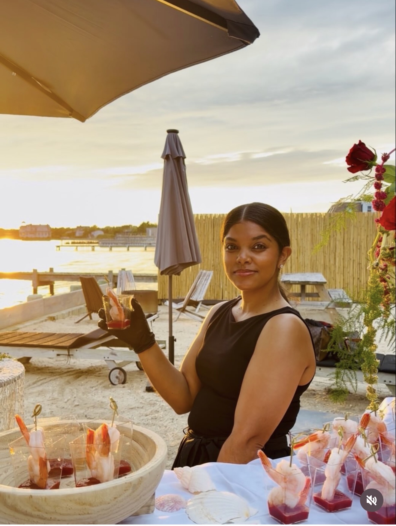 Nadia in black beside a waterfront raw bar of shrimp cocktail servings at golden hour