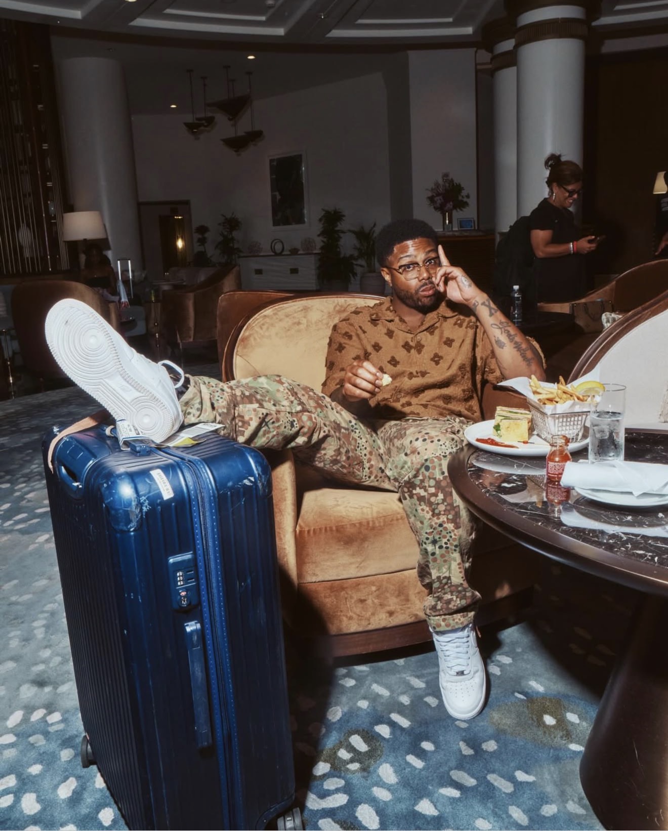 Kwame Onwuachi seated in a hotel lobby with a club sandwich, fries and a suitcase beside him